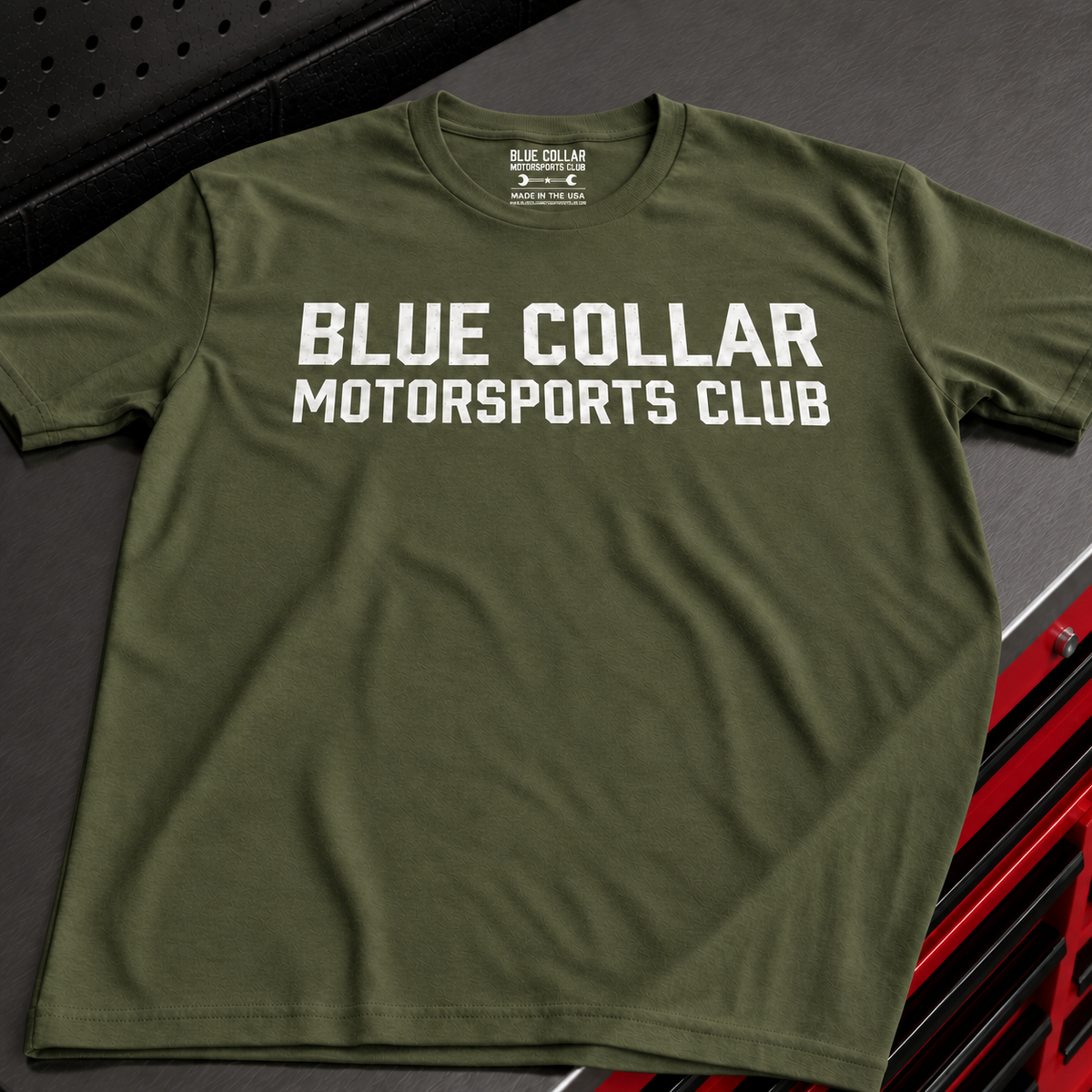 An olive green crewneck t-shirt with 'BLUE COLLAR MOTORSPORTS CLUB' printed in bold white letters across the chest, displayed on a dark textured surface.
