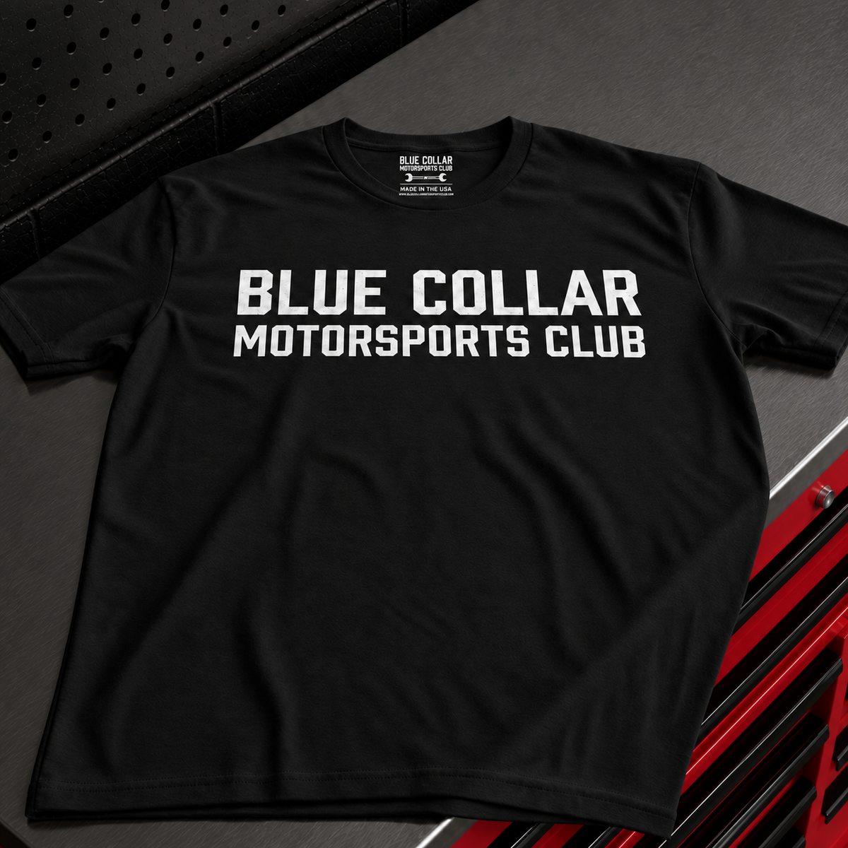 A black short-sleeve t-shirt with 'BLUE COLLAR MOTORSPORTS CLUB' printed in bold white capital letters across the chest, displayed on a dark textured background with red and black diagonal graphic elements in the corner.