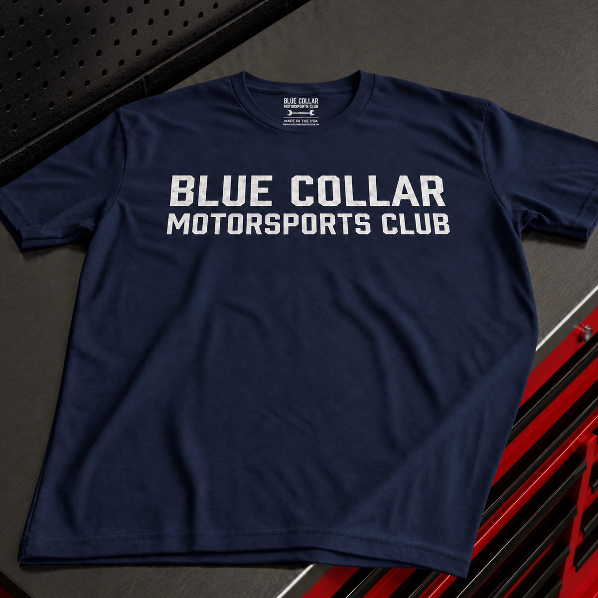 A navy blue short-sleeve t-shirt with 'BLUE COLLAR MOTORSPORTS CLUB' printed in bold white letters across the chest, laid flat on a dark textured surface with red and black automotive-themed props.