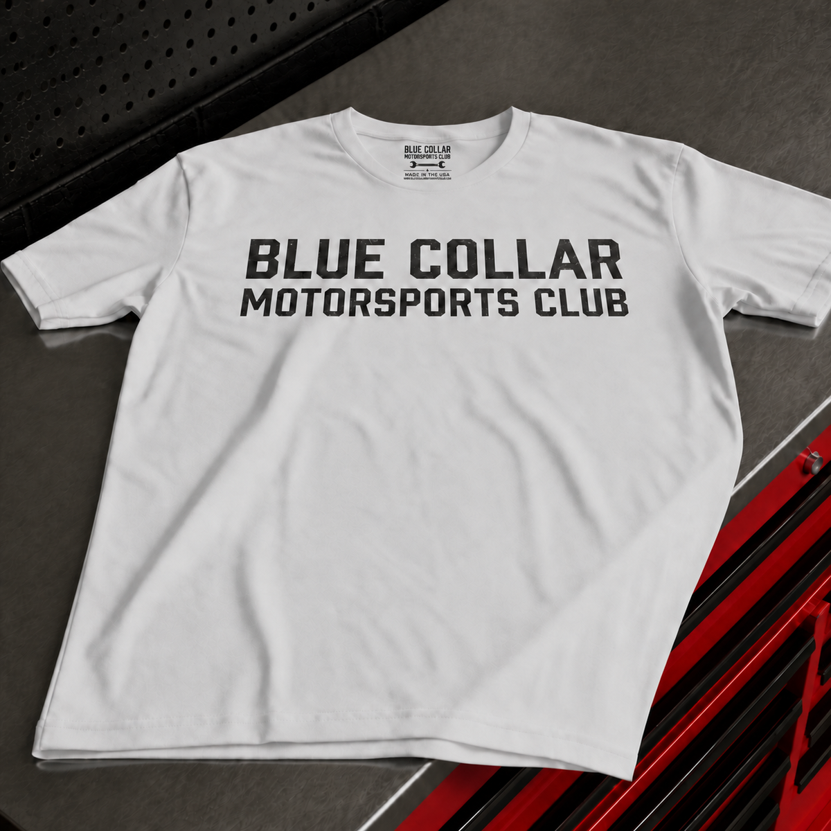 A white short-sleeve t-shirt with 'BLUE COLLAR MOTORSPORTS CLUB' printed in bold black letters across the chest, laid flat on a dark gray textured surface with red and black vehicle graphics in the corner.