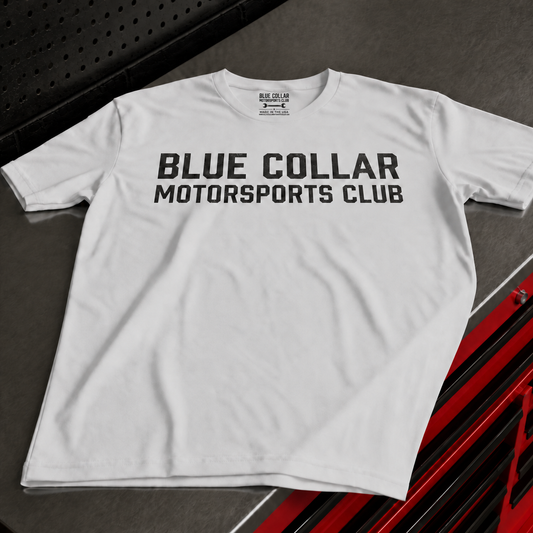 A white short-sleeve t-shirt with 'BLUE COLLAR MOTORSPORTS CLUB' printed in bold black letters across the chest, laid flat on a dark gray textured surface with red and black vehicle graphics in the corner.